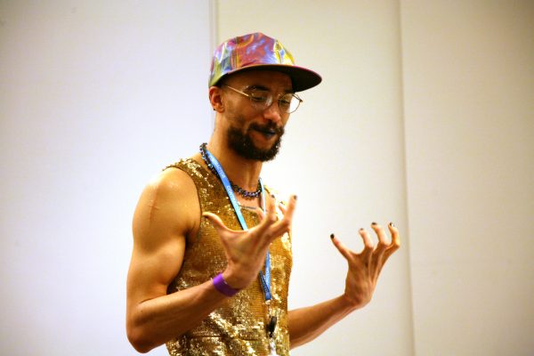 Photo of a young person with a beard and well-toned biceps., who is wearing a gold tank top, a blue necklace, a pair of round stylish glasses, and a holographic hat. They are explaining something using hand gestures.