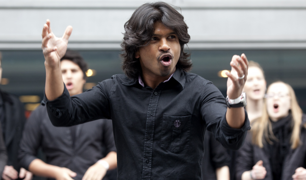 Photo of a young man leading the crowd behind him to sing. He wears a black shirt and is confidently conducting with hand gestures.