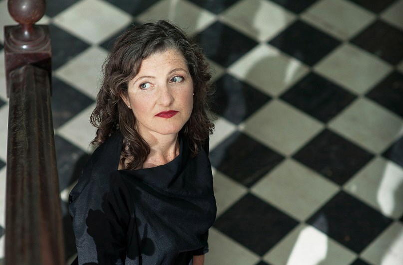 Photo of a white woman with simple makeup and curly hair. Her dark dress and the black-and-white chessboard-patterned floor behind her creates a mysterious atmosphere.