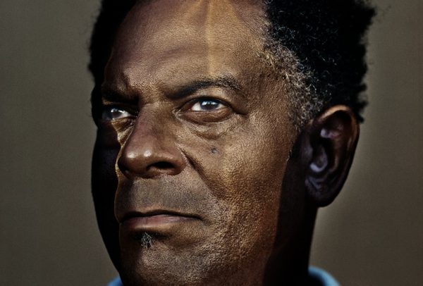 A closeup photo of a Black man gazing into the distance. His face is partially lit by a golden reflection.