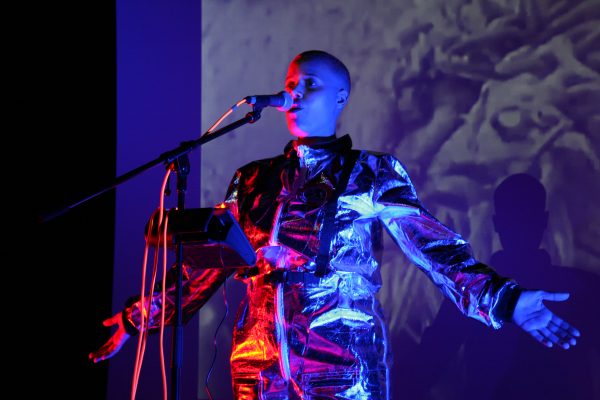 Photo of a performing artist standing behind a mic, who is lit by red and blue side lighting. Their arms are spread to the sides and their palm facing forward.
