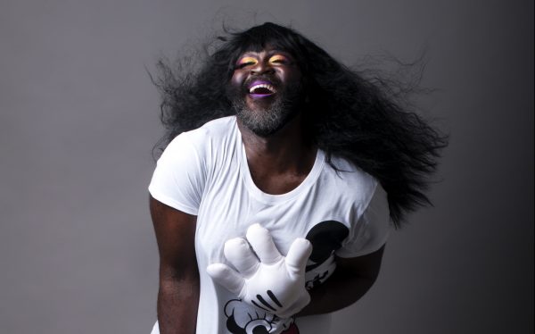 A Black person with bright yellow and purple makeup and a beard laughs glamorously in joy, their lustrous long hair sways to the back. They are wearing a Mickey Mouse T-shirt and a Mickey Mouse glove.