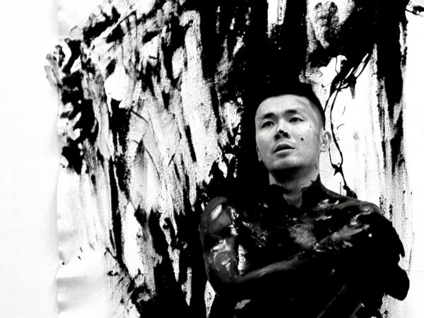 A black-and-white photo of an Asian young man standing in front of a large unstretched canvas overed in marks of paint. He has paint marks over his nose. His body is covered in paint from the neck down. He gazes into the distance with a serious expression.