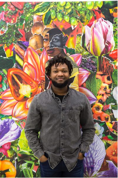 Larry, a young man with short beard dresssed in a grey shirt, smiles at the camera. Behind him is one of his artworks, a colourful collage of people, plants and fruits