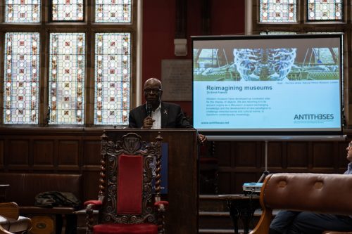 Black man in black suit with a airplane-shaped pin speaking into a hand-held mic. The powerpoint behind him says: Reimagining Museums