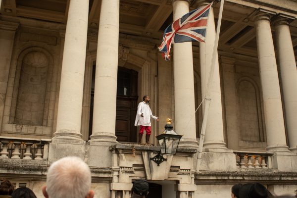 Photo of a Black actor with red victorian costume standing under a UK flag at Old Royal Naval College, the audiences watch from below