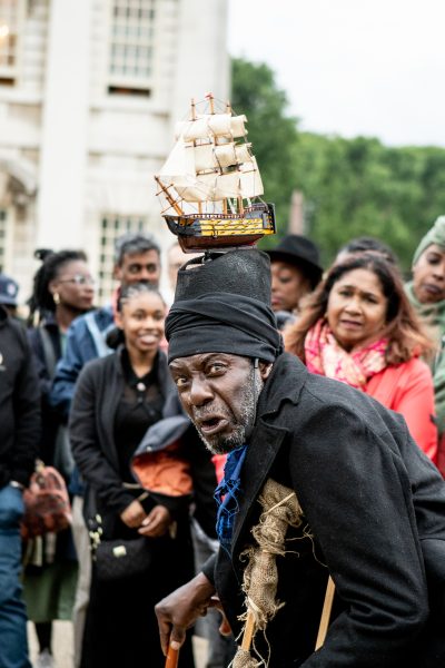 A Black man in wood and rug crutches wearing a black hat with a model sailing ship on top