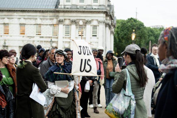 A woman wearing a hat, a tartan scarf and dress holding a fabric sign that says 'JOIN US'