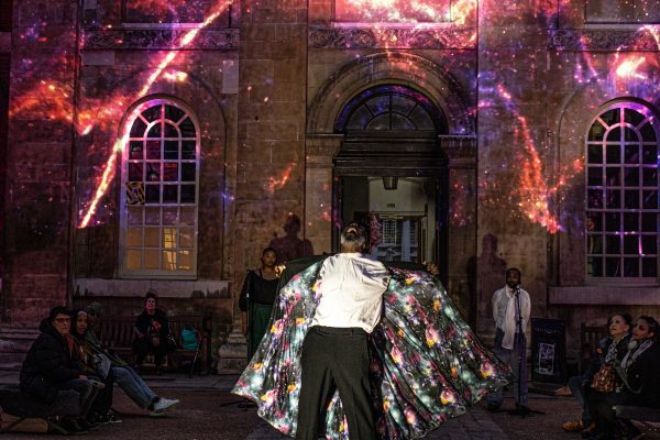 A Black man in a white shirt and black pants standing in front of a building with a large light show, the cosmic prints in his jacket blends in with the nebular projected behind him.