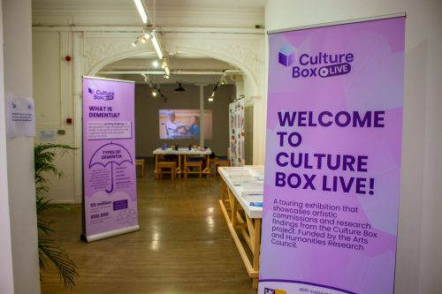 Image shows an exhibition space with large craft tables and a projection. There are two purple standing banners saying 'welcome to Culture Box Live, a touring exhibition that showcases artistic commissions and research findings from the Culture Box project. Funded by the AHRC', and 'What is Dementia?' respectively.